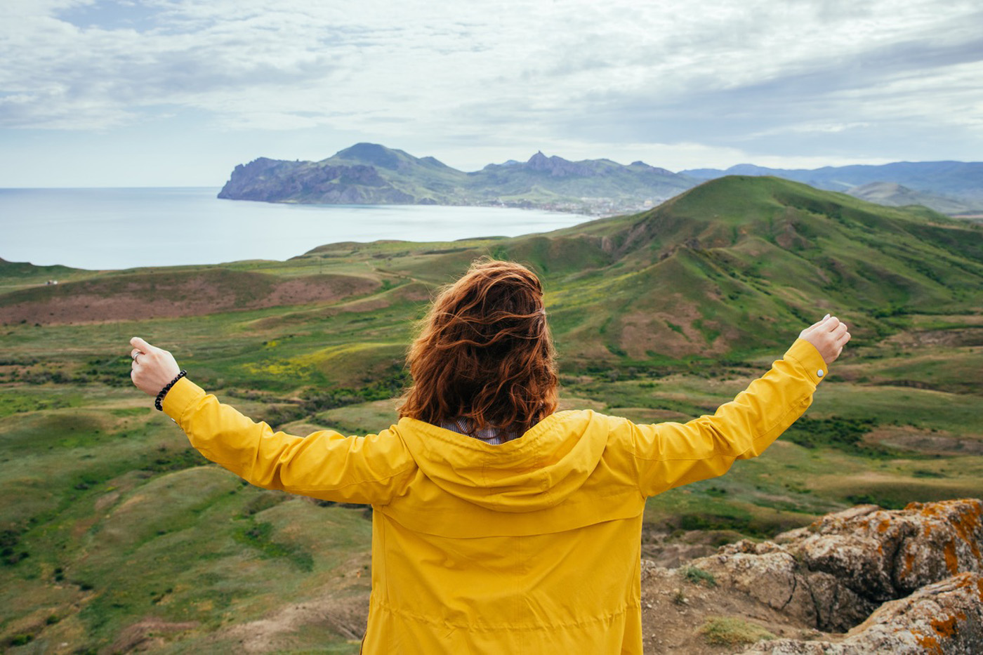 woman in raincoat standing above hills and water with arms open