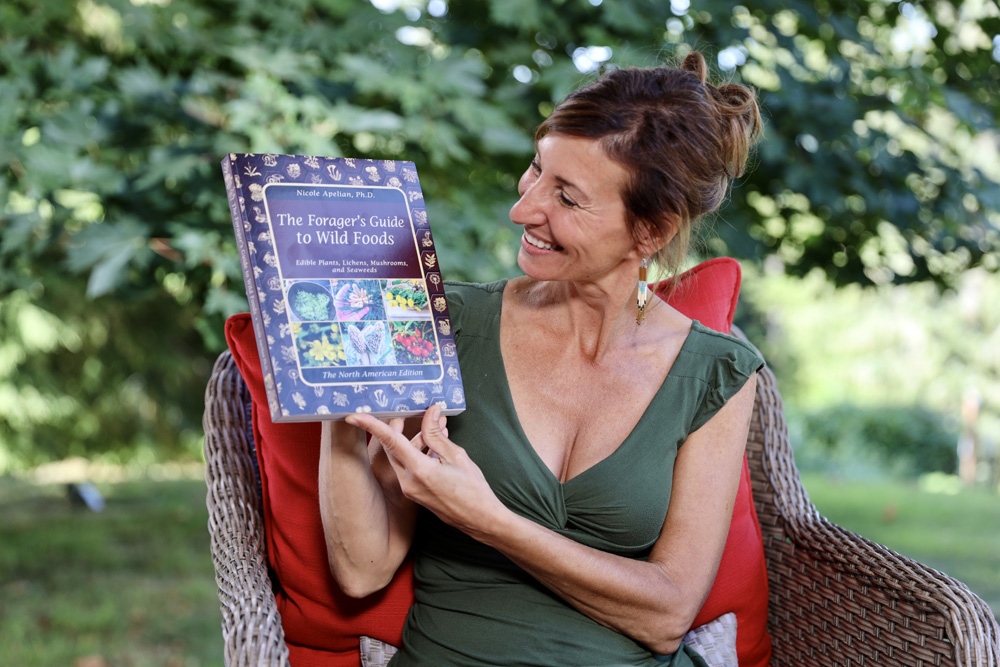 Nicole Apelian sitting on wicker chair holding her Wild Foods book